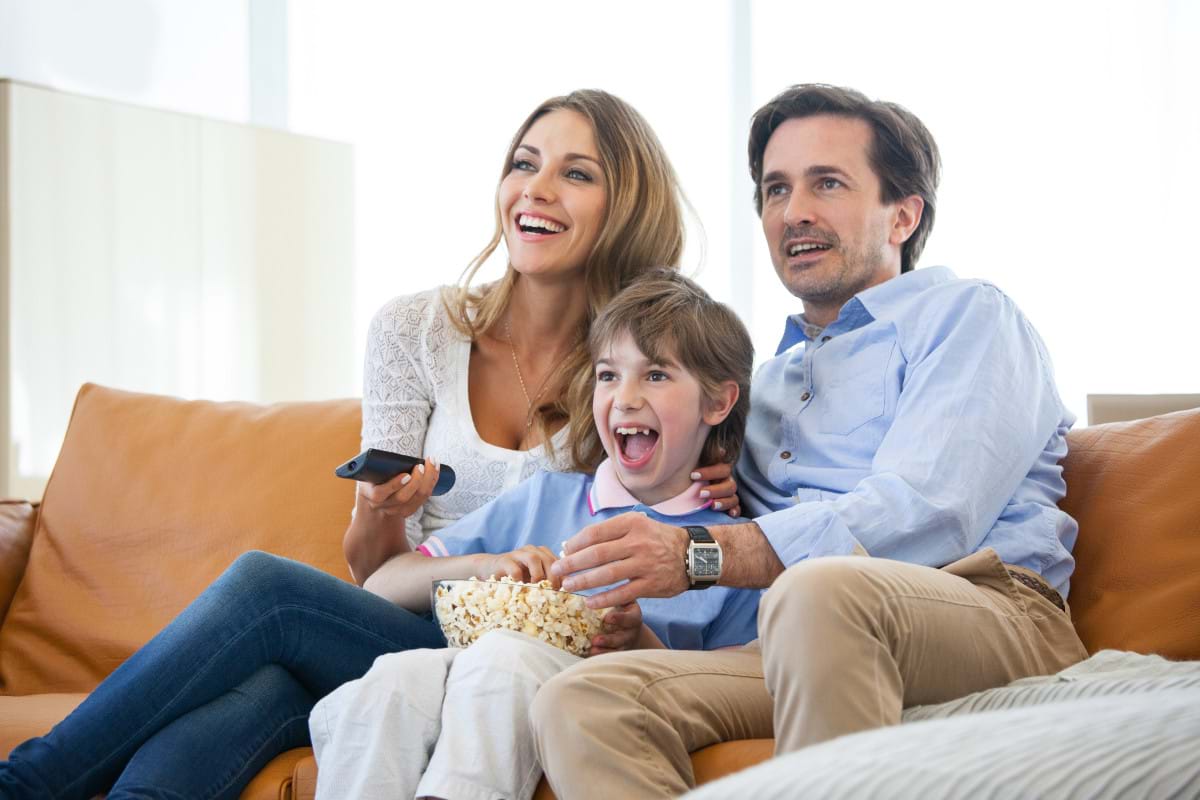 Smiling parents and son watching tv and eating popcorn while sitting on couch